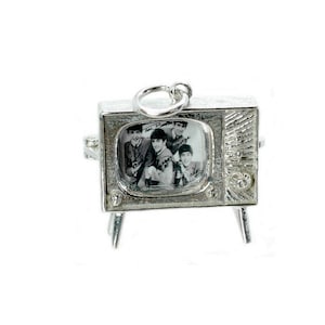 May include: A silver-toned charm in the shape of a vintage television set. The screen displays a black and white image of a band. The charm has a loop at the top for attachment and rests on four small legs.