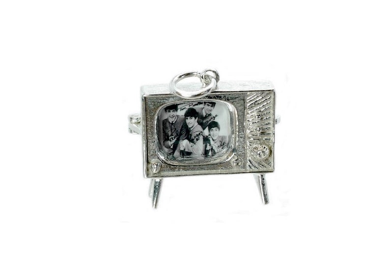 May include: A silver-toned charm in the shape of a vintage television set. The screen displays a black and white image of a band. The charm has a loop at the top for attachment and rests on four small legs.