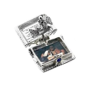 May include: A silver-toned, hinged pendant with an open rectangular compartment revealing a small image of a religious scene. The pendant has ornate detailing, including a small blue gem and a decorative top with text.