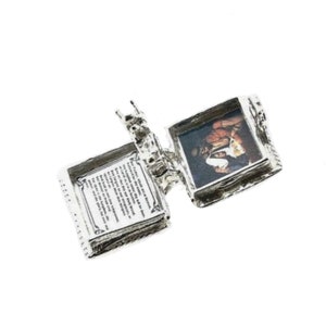 May include: A silver-colored, hinged locket with an open book design. One side displays a small image of a person, while the other side features printed text. The locket is set against a white background.