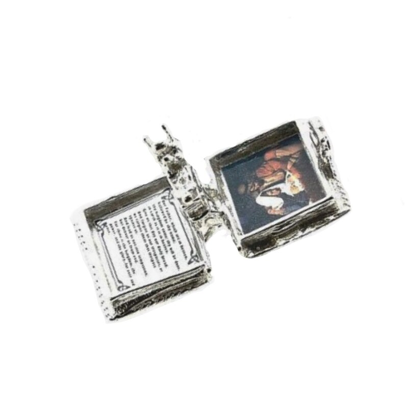 May include: A silver-colored, hinged locket with an open book design. One side displays a small image of a person, while the other side features printed text. The locket is set against a white background.