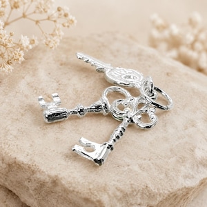 May include: A set of three small, shiny silver-toned metal key charms linked together by a circular jump ring. Each key features a distinct ornate head design with intricate cutouts and a long, slender shaft ending in a notched bit.