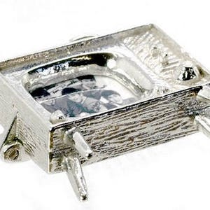 May include: A silver-colored miniature television charm with a textured surface. The screen displays a black and white image of a group of people. The charm has four legs and a small antenna.