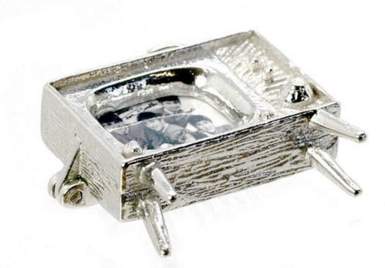 May include: A silver-colored miniature television charm with a textured surface. The screen displays a black and white image of a group of people. The charm has four legs and a small antenna.