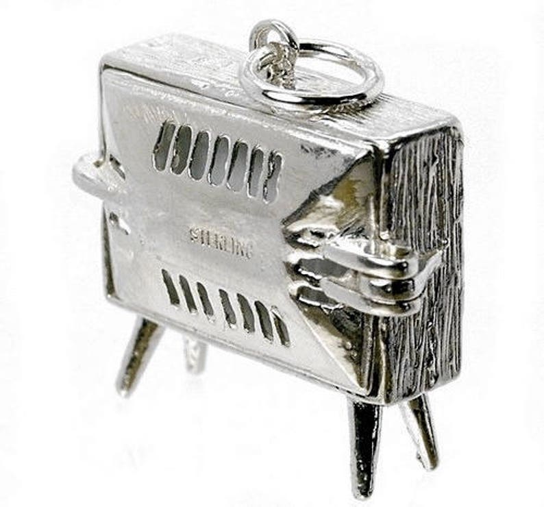 May include: A detailed, silver-toned charm in the shape of a vintage television set. The charm has a textured surface, vent-like openings, and small legs. The word "STERLING" is engraved on the front. A small ring is attached at the top.