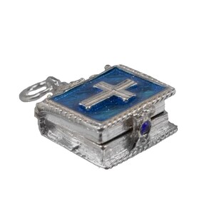 May include: A silver-colored miniature book pendant with a blue enamel cover featuring a silver cross. The pendant has a small loop for a chain and a blue gemstone on the side. The edges are detailed with a rope-like design.