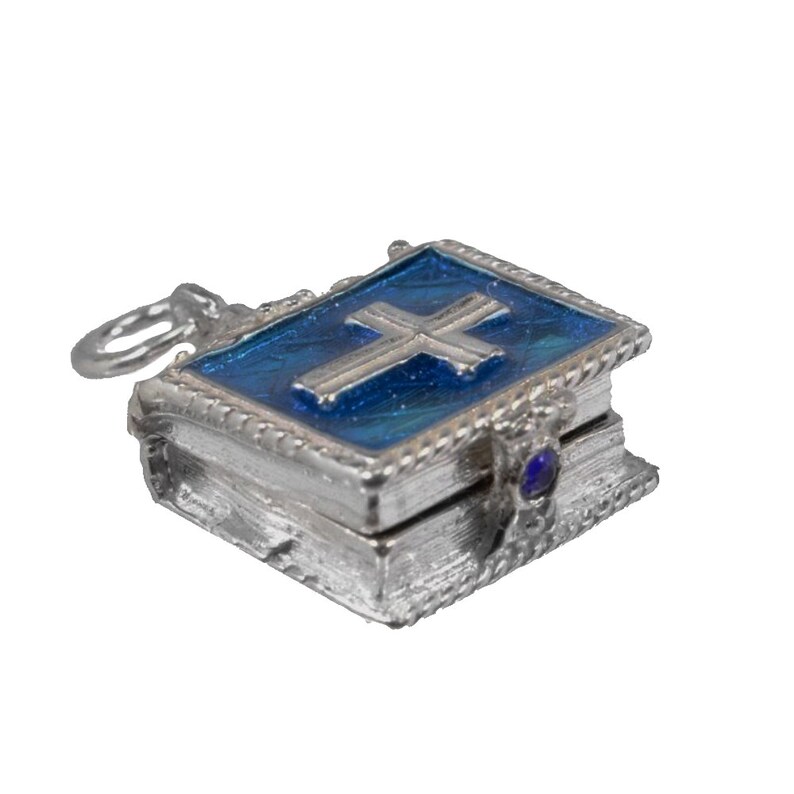 May include: A silver-colored miniature book pendant with a blue enamel cover featuring a silver cross. The pendant has a small loop for a chain and a blue gemstone on the side. The edges are detailed with a rope-like design.