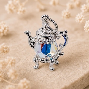 May include: A silver-toned metal teapot charm featuring an intricate, ornate lid and handle design. The body of the teapot is crafted from a faceted, clear crystal that displays internal refractions of blue, red, and yellow light. A jump ring is attached.