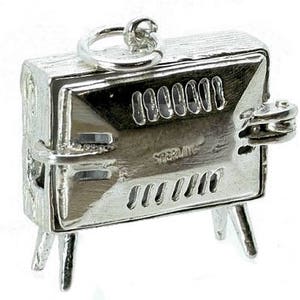 May include: A small, silver-colored charm in the shape of a barbecue grill. The charm has a rectangular body with a hinged lid, a handle, and three small legs. The word "STERLING" is visible on the front.