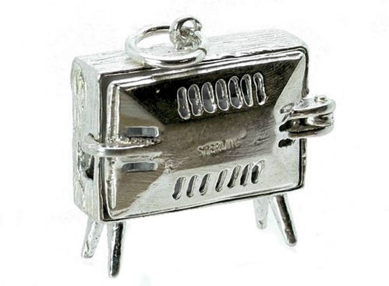 May include: A small, silver-colored charm in the shape of a barbecue grill. The charm has a rectangular body with a hinged lid, a handle, and three small legs. The word "STERLING" is visible on the front.