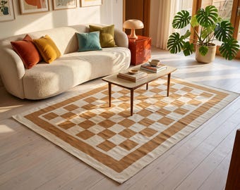 May include: A rectangular area rug featuring a repeating checkerboard pattern in alternating shades of tan and cream. The rug has a solid tan border framing the central grid design, laid flat on a light-colored wooden floor in a living room.
