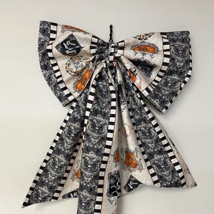 May include: A large fabric bow featuring a cream, black, and orange pattern with black and white striped borders. The fabric displays ornate damask-style motifs, skulls, and text labels including Gone Batty and Trick or Treat.