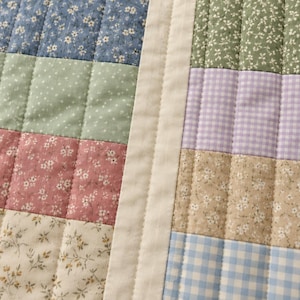 May include: A quilted fabric textile featuring a patchwork design of rectangular panels. The pattern includes blue, green, pink, and cream floral prints alongside lavender and blue gingham checks, all separated by a solid cream border.