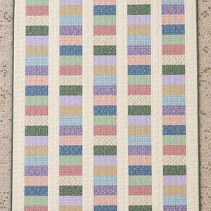 May include: A rectangular quilt featuring five vertical columns of alternating fabric rectangles in various pastel and muted colors, including blue, green, pink, purple, and tan, with floral and plaid patterns, all bordered by a solid cream fabric.