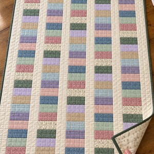 May include: A rectangular quilted table runner featuring five vertical columns of small, multi-colored fabric rectangles in shades of blue, pink, green, purple, and tan, separated by cream-colored sashing and finished with a dark green border.