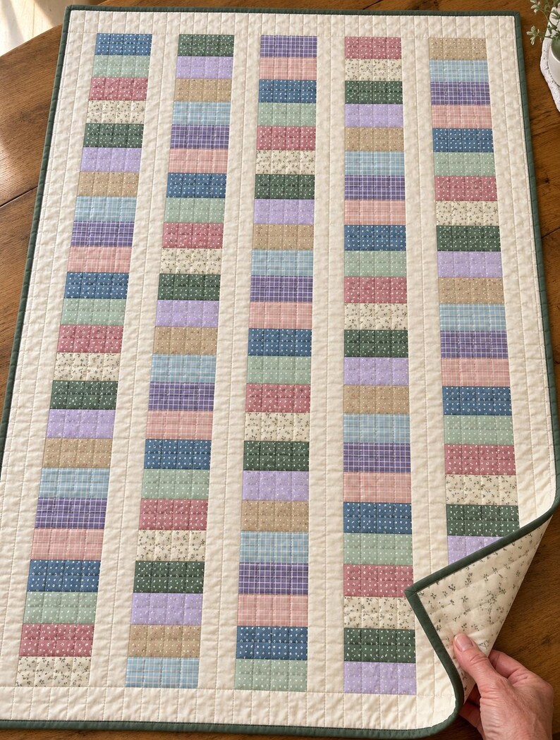 May include: A rectangular quilted table runner featuring five vertical columns of small, multi-colored fabric rectangles in shades of blue, pink, green, purple, and tan, separated by cream-colored sashing and finished with a dark green border.