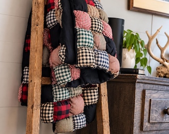 May include: A handmade puff quilt featuring a patchwork design of individual fabric squares in red, black, green, and tan gingham and buffalo check patterns, draped over a tall, weathered wooden ladder with a rustic, unfinished texture.