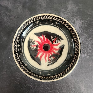 May include: A round ceramic dish featuring a central red sun graphic surrounded by black hand silhouettes. The border is decorated with a black and white rope pattern and small star icons against a cream-colored background.