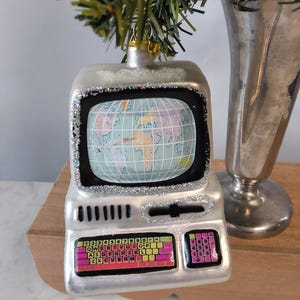 May include: A silver computer-shaped Christmas ornament featuring a screen displaying a world map. The keyboard area has colorful keys with letters and numbers, and a separate numeric keypad. The frame is accented with silver glitter.