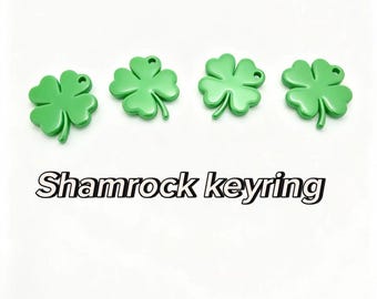 May include: Four bright green four-leaf clover charms, each featuring a small hole at the top for attachment. The text Shamrock keyring is displayed in a bold, black-outlined font below the four clover-shaped metal pieces.