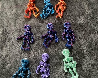 May include: A collection of nine articulated skeleton figurines in various colors including orange, blue, purple, and light green. Each small figure features a detailed skull, rib cage, and jointed limbs, arranged in a loose formation.