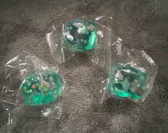 May include: Three individual translucent teal resin pieces, each encased in clear plastic packaging. The resin shapes contain embedded holographic star-shaped glitter and small white seashell-like decorative elements throughout.