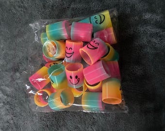 May include: A collection of small, cylindrical rainbow-colored plastic springs packaged in a clear plastic bag. Each spring features a gradient of pink, yellow, and blue hues, with some displaying a black printed smiley face icon on the side.