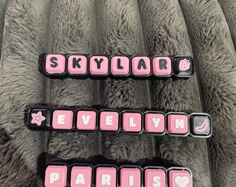 May include: Three black rectangular hair clips featuring pink square tiles with black letters. The top clip spells SKYLAR with a strawberry icon. The middle clip spells EVELYN with a star and banana icon. The bottom clip spells PARIS with a heart icon.