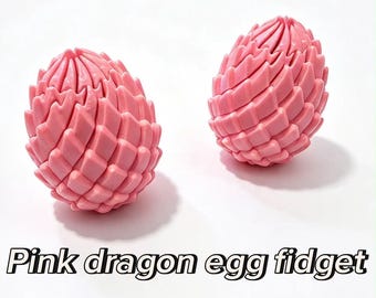 May include: Two pink dragon egg fidget toys with a textured, scale-like surface. The objects are oval-shaped and feature a repeating geometric pattern. The text "Pink dragon egg fidget" is displayed in black letters below the two items.