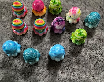 May include: A collection of eleven small octopus figurines featuring various patterns and colors, including rainbow stripes, pink tie-dye, green camouflage, purple galaxy, and blue checkered designs, each with a smooth, rounded head and tentacles.