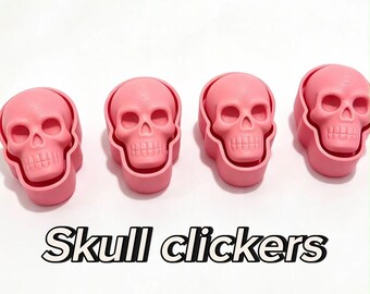 May include: Four pink skull clickers arranged in a horizontal row. Each item features a detailed skull design with hollow eye sockets and a defined jawline. The text "Skull clickers" is printed in black letters below the four pink objects.