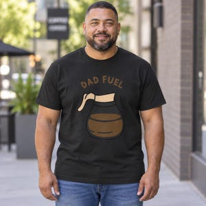 Può includere: Una t-shirt nera a maniche corte con la grafica di una caffettiera in vetro riempita di liquido marrone. Sopra la caffettiera, il testo DAD FUEL è stampato in un carattere marrone curvo. La maglietta ha un girocollo e una vestibilità standard.