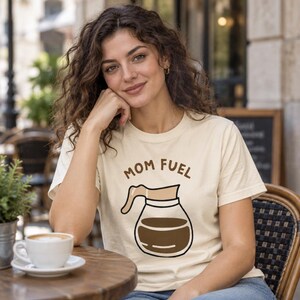 Puede incluir: Una camiseta de manga corta color crema con un gráfico de una jarra de café de cristal llena de líquido marrón oscuro. Sobre la jarra, el texto MOM FUEL aparece escrito en letras mayúsculas de color marrón.