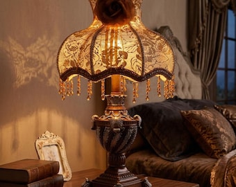 May include: A table lamp with a dark brown, ornate base and a bell-shaped fabric shade featuring a floral lace pattern. The shade is topped with a fabric rose and trimmed with hanging amber-colored crystal beads that glow when lit.