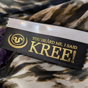 以下が含まれることがあります： 黒と金のリボンに「YOU HEARD ME, I SAID KREE!」の文字と金のシンボルが書かれています。