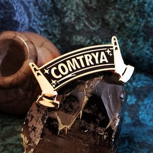 以下が含まれることがあります： 黒いバナーに「COMTRYA」と書かれた、金色のエナメルピン。バナーの両側に金色の手が2つある。