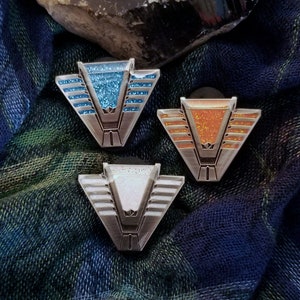 Peut inclure: Trois épingles en émail avec un motif géométrique et des accents de paillettes. Les épingles sont en bleu, orange et blanc.