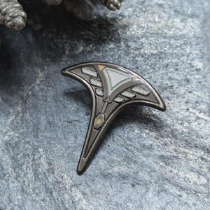 May include: A metallic enamel pin featuring a dark gunmetal finish with a curved, fan-like top section and a tapered base. The design includes geometric cutouts filled with light gray enamel and small gold-colored accents along the edges.