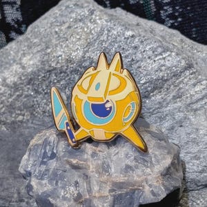 Peut inclure: Une broche en émail ronde, jaune et crème, représentant un personnage stylisé avec un grand œil bleu et un petit couteau bleu tenu dans une main. Le personnage a des oreilles pointues et un corps circulaire avec des contours en métal doré.