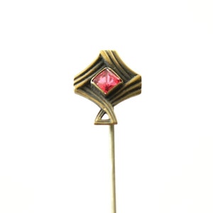 May include: A gold-toned pin with a geometric design and a red rhinestone in the center.