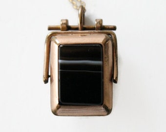 May include: A gold-toned locket pendant with a black and white agate stone. The locket has a hinged opening and a decorative design.