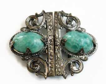 May include: A two-piece metal cloak clasp featuring an ornate, dark silver-toned filigree design with scrolling patterns. Each side is centered with a large, oval-shaped, textured turquoise-green stone held in place by four small metal prongs.