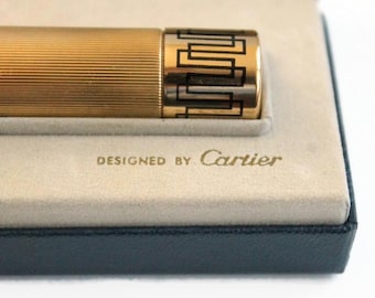 May include: A gold-toned lighter with a ribbed body and a geometric black design on the end. The lighter is in a cream-colored case with the words "DESIGNED BY Cartier" in gold lettering.