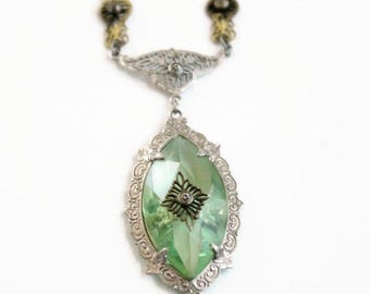 May include: A necklace featuring a large, oval, light green faceted stone set in an ornate silver-toned metal frame. The pendant is suspended from a chain with decorative silver and gold-toned metal links, showcasing intricate filigree detailing.