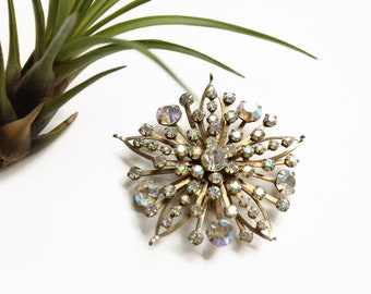 May include: A gold-toned brooch with a starburst design, featuring many clear iridescent rhinestones.