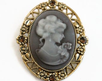 May include: An oval brooch featuring a grey cameo of a profile portrait set within a gold-toned metal frame. The frame is decorated with small floral motifs and embedded with tiny brown rhinestones at the top, bottom, and sides.