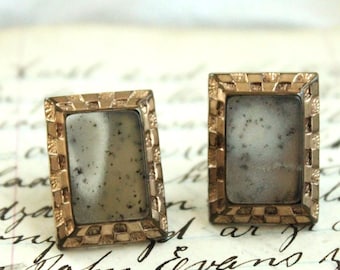 May include: A pair of rectangular cufflinks featuring a metallic gold-toned frame with a textured, notched border. The center of each cufflink contains a smooth, light gray stone inlay with subtle dark speckles throughout the surface.
