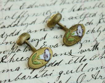 May include: A pair of oval-shaped brass cufflinks featuring a hand-painted floral design with white and purple petals and green leaves. The cufflinks have a metallic finish and a classic toggle-style backing mechanism for securing to a shirt cuff.