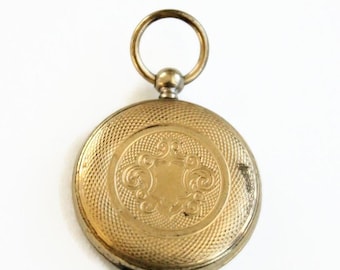 May include: A gold-colored pocket watch with a textured, patterned surface. The watch features an ornate, engraved design in the center and a small loop at the top for attaching a chain. The watch is round and appears to be vintage.