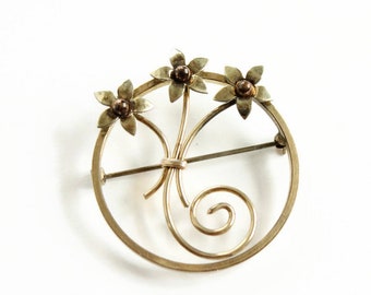 May include: A gold-toned brooch with three stylized flower designs. The flowers have brown centers and are set within a circular frame.